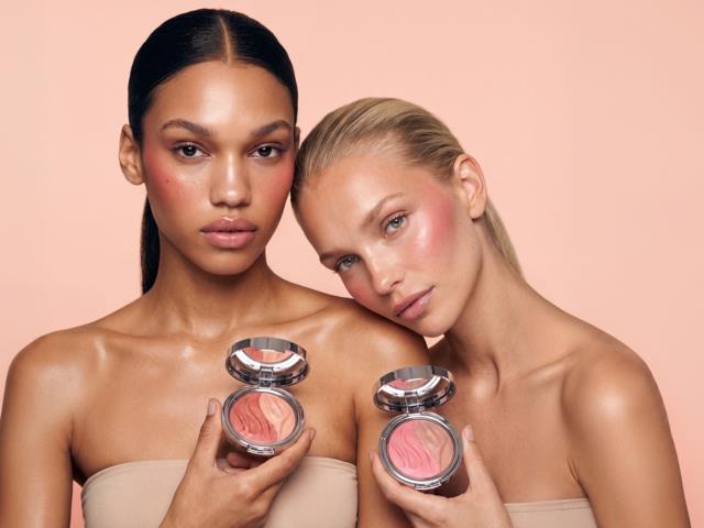 Two women hold open blush products in front of them, displaying their makeup and glowing skin against a pink background.