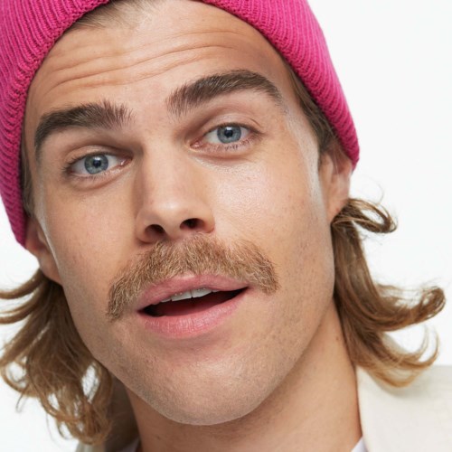 En ung man med långt hår, en rosa mössa, blå ögon och mustasch ser förvånad eller nyfiken ut.