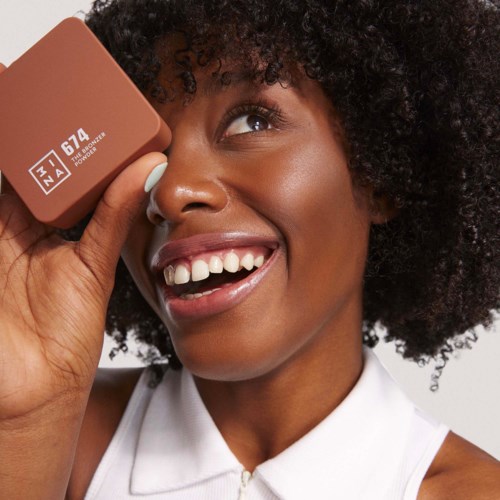 A smiling woman with curly hair holds a makeup product near her eye, showing bright teeth and joy.