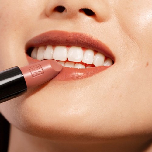 A person smiles while applying nude lipstick to their lips.