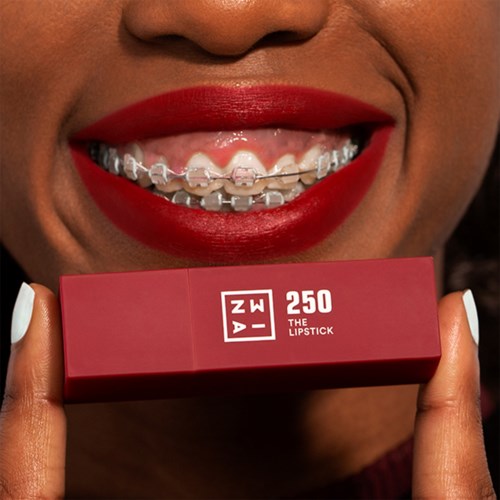A person showing teeth with braces, holding a red lipstick box near their mouth.