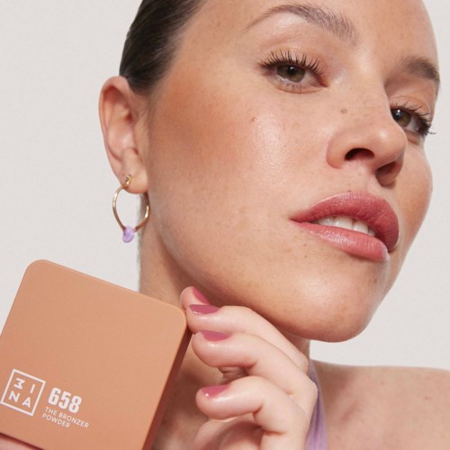 A woman with light makeup and earrings holds a beige box labeled '3INA 658.'