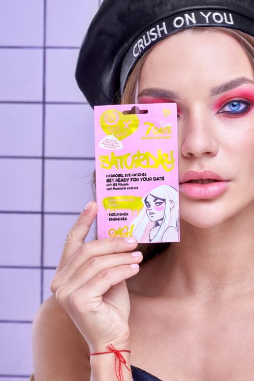En kvinne med lyst sminke holder en rosa pakke merket 'Hydrogel Eye Patches' nær ansiktet sitt. Hun har på seg en svart hatt som sier 'Crush on You.'