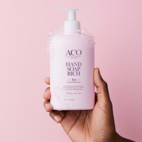 Een hand houdt een roze fles met 'ACQ Hand Soap Rich' en een pomp in de hand voor een roze achtergrond.