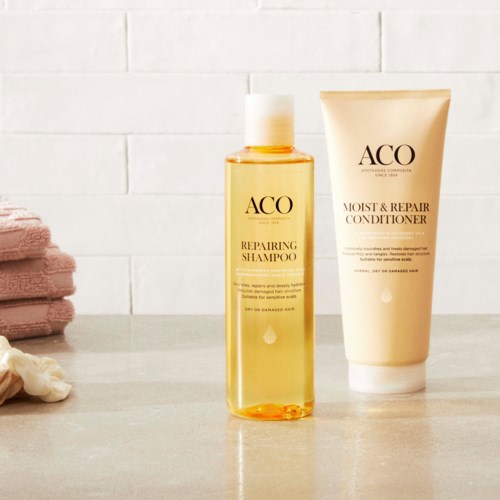 Two hair care products, shampoo and conditioner, are placed on a bathroom counter near towels and a white tiled wall.