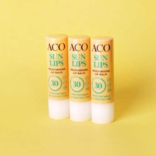 Kolme ACO Sun Lips kosteusvoidetta huultenhoitoaineita SPF 30, taustalla keltainen.