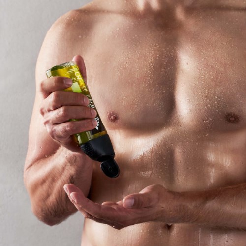 En naken man smörjer in kroppsolja eller lotion på bröstet mot en ljus bakgrund.