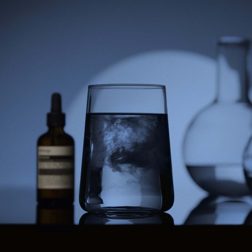 Ett glas fyllt med vatten med reflektion och några laboratorieflaskor i bakgrunden.