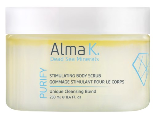 En gul krem i en gjennomsiktig krukke merket "Alma K. Dead Sea Minerals" for kroppsskrubb, med blå tekst og et geometrisk mønster.