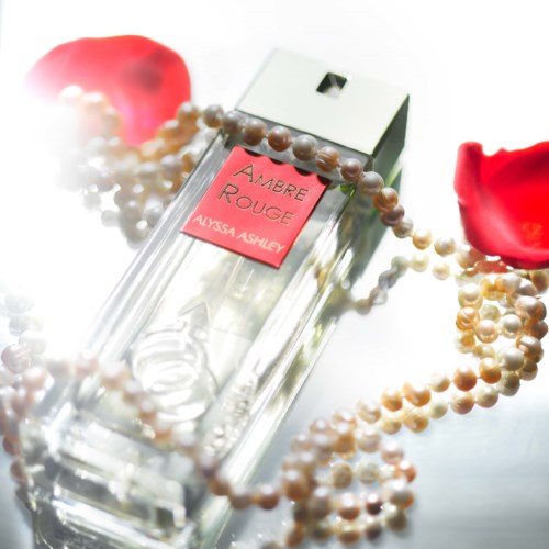 Een heldere parfumfles met het label 'Amber Rouge' door Alyssa Ashley, omgeven door parelkettinkjes en rozenblaadjes.