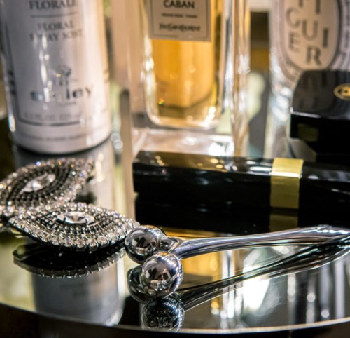 Zbiór produktów do pielęgnacji i biżuterii, w tym perfumy, naszyjnik i lustro, na błyszczącej powierzchni.