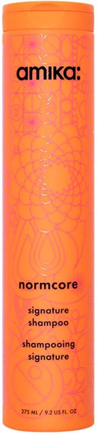 Pitkä oranssi shampoopullo, jossa on mustaa ja vaaleanpunaista tekstiä ja abstrakteja kuvia. Se lukee 'Amika: Normcore Signature Shampoo.'