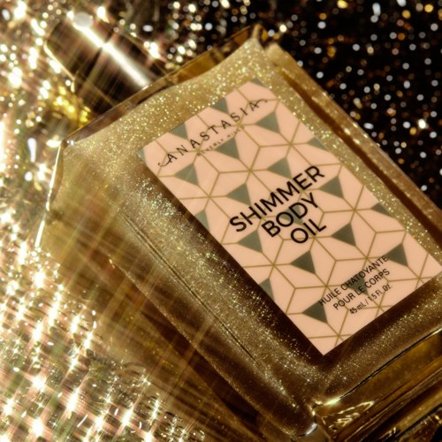 A gold glitter box contains a bottle of shimmer body oil on a sparkling surface.