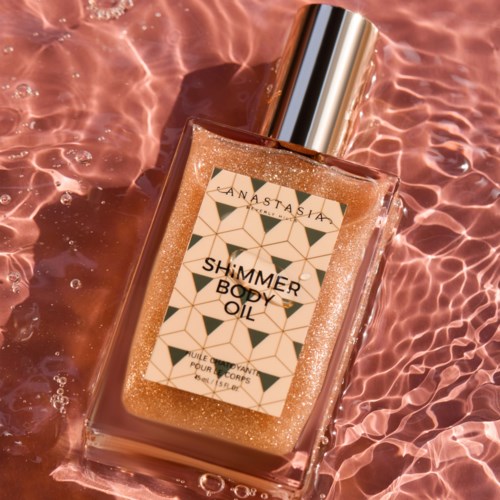 A bottle of shimmer body oil lying on water, with sunlight reflecting on it and a shiny silver cap.