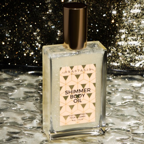 A clear glass bottle with a golden cap, labeled 'Shimmer Body Oil' by Anastasia, on a reflective silver background.