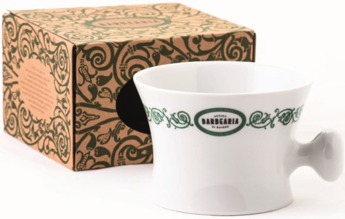 En vit kaffekopp med handtag och en grön och svart logga, bredvid en dekorativ låda med ett virvlande mönster.