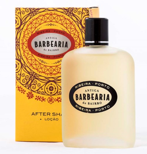 Een witte fles met zwarte dop, gelabeld "Antiga Barbearia de Bairro". Doos achter is geel en oranje met decoratieve patronen.