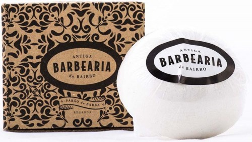 Een witte scheerzeep in een ronde container naast een decoratieve bruine en zwarte doos, beide gelabeld 'Barbearia' en 'Antiga'.