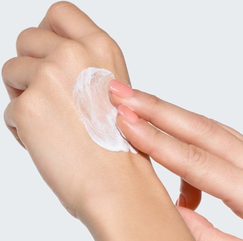 Person som applicerar vit kräm på handryggen med fingrarna. Bakgrunden är ljusgrå.