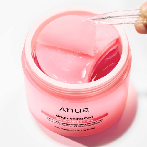 Ein rosa Behälter mit Anua Brightening Pad und einem transparenten Spatel, der einen Pad hebt.