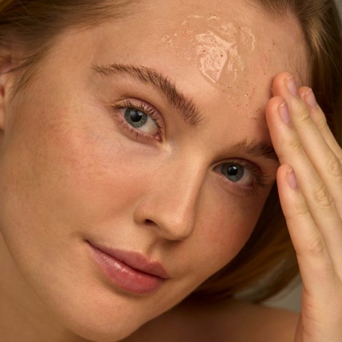 A woman applies a facial mask on her forehead, with clear, smooth skin and light hair. She gently touches her face.