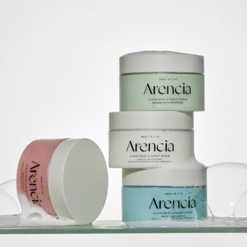 Gestapelte Gläser von Arencia Kopfhaut- und Körperscrub in Pastellfarben mit einfachen Textetiketten. Offene Dose mit rosa Peeling.