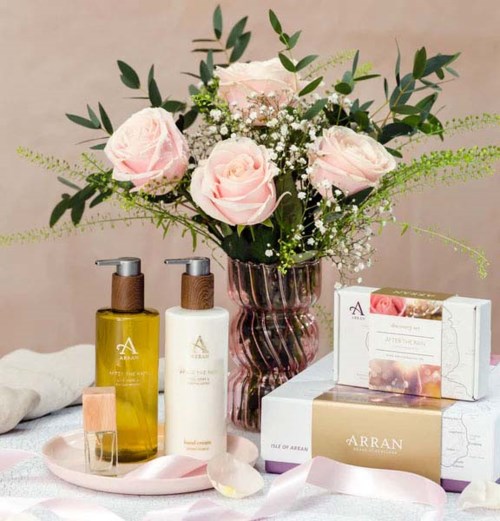 A bouquet of pink roses and white baby's breath in a pink glass vase, skincare products, and a box on a table.