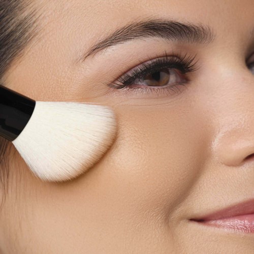 A woman applies makeup with a brush on her cheek, smiling gently with smooth, clear skin and well-groomed eyebrows.