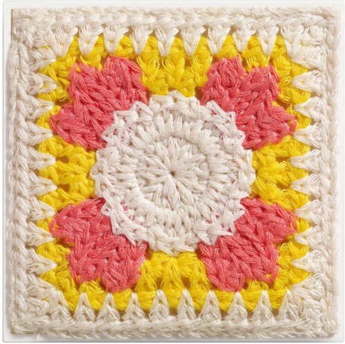 Et fargerikt heklet kvadrat med hvit sentrum, rosa blomstermønster, gul bakgrunn og hvit kant.