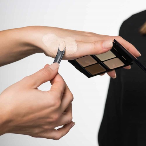 En person applicerar ljus foundation eller concealer på armen med en liten borste, nära en ögonskuggspalett.