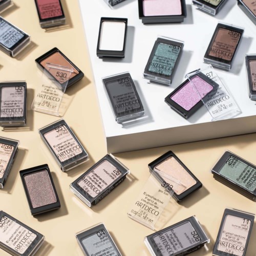 En udstilling af farverige makeup-pletter og øjenskygger i rektangulære beholdere, arrangeret på beige og hvide overflader.