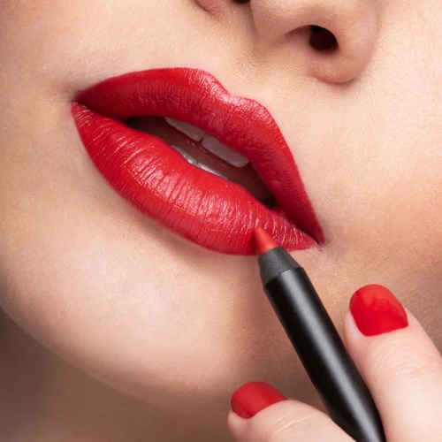 A woman applies bright red lipstick on her lips, with her finger holding the lipstick pencil against her bottom lip.