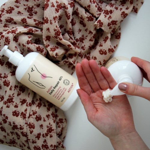 En person bruker lotion på hånden med et blomstrete stoff og en hudpleieflesk i nærheten.