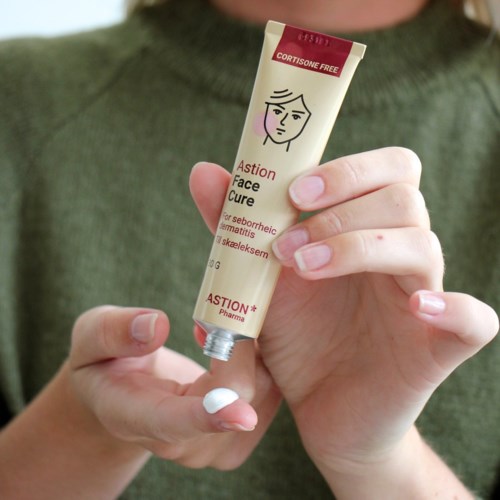 En person håller i en tub ansiktskräm märkt 'Astion Face Cure' framför en grön topp. Rör är rött och beige.