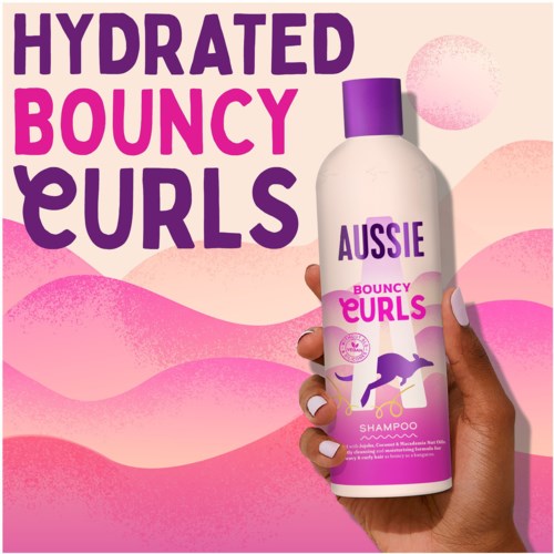 En hand håller en flaska Aussie Bouncy Curls-schampo med lila lock, mot en bakgrund av rosa och persikofärgade vågiga former.