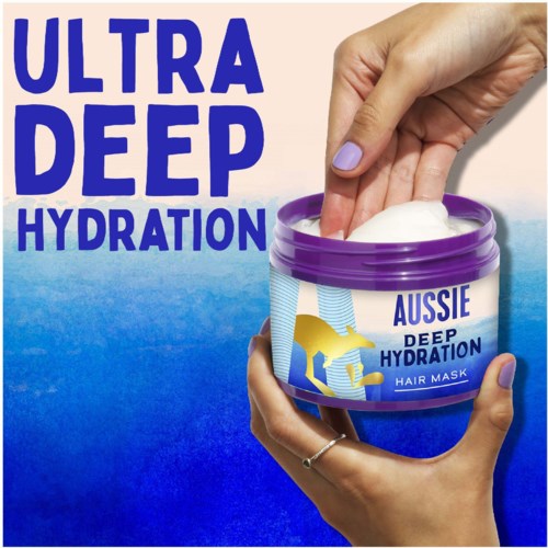 Käsivarsi laittaa valkoisen voiteen purkkiin, jossa lukee 'Aussie Deep Hydration Hair Mask' ja taustalla sininen.