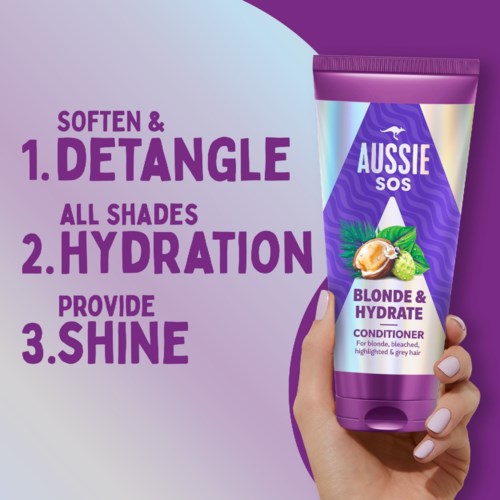 En hand håller en lila flaska Aussie hårbalsam med text som lyfter fram fördelar som att reda ut, återfukta och ge glans.