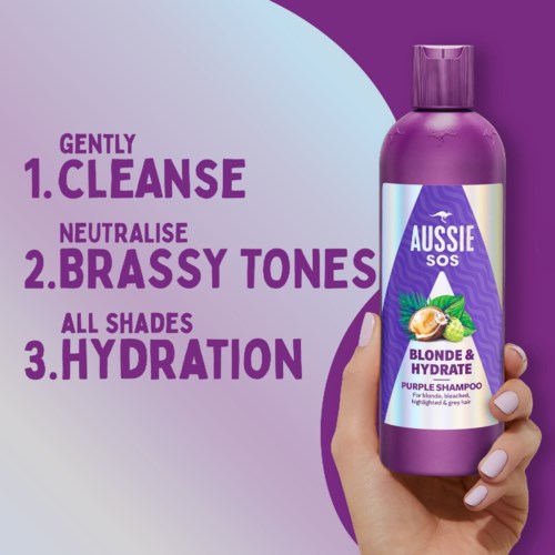 Violetin värinen shampoopullo, jossa lukee 'Aussie SOS Blonde & Hydrate,' kädessä, edistää hiusten hyvää kuntoa.