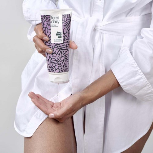 En kvinna i en vit morgonrock använder en tub Femí daily tea tree oil på sin hud.