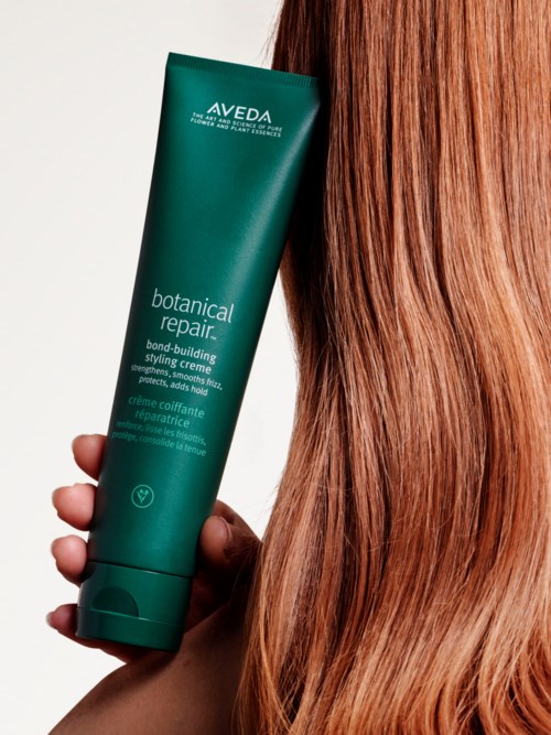 Henkilö pitää vihreää Aveda Botanical Repair -tuotepulloa pitkän, suoran, punertavan hiuksen vieressä.