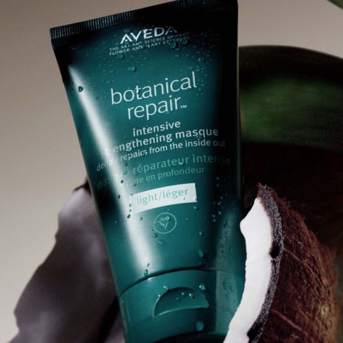 Tuba koloru turkusowego Aveda botanical repair maski do włosów z kroplami wody, trzymana przez rękę na ciemnym tle.