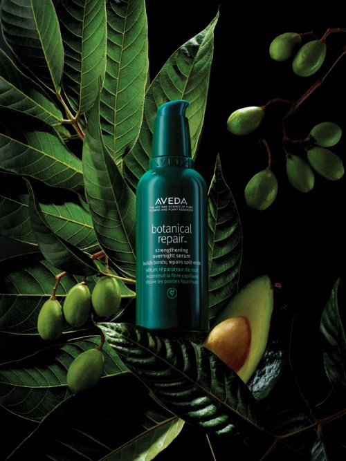 Sininen pullo Aveda botanical repair -hiustuotteesta vihreiden lehtien ja hedelmien ympäröimänä mustalla taustalla.