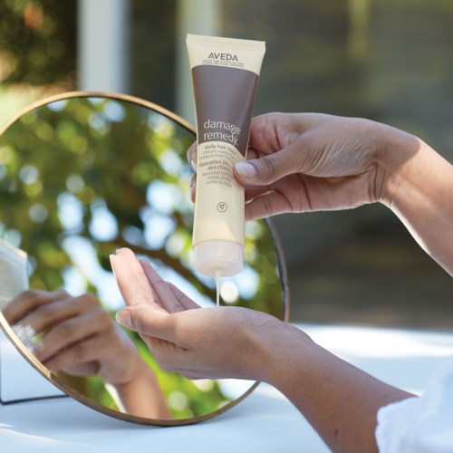 En person applicerar Aveda damage remedy hårbehandling framför en spegel utomhus.