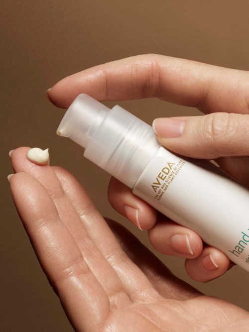 Iemand brengt Aveda handcrème aan met een pomp. De achtergrond is simpel. Focus op handen en de crème.