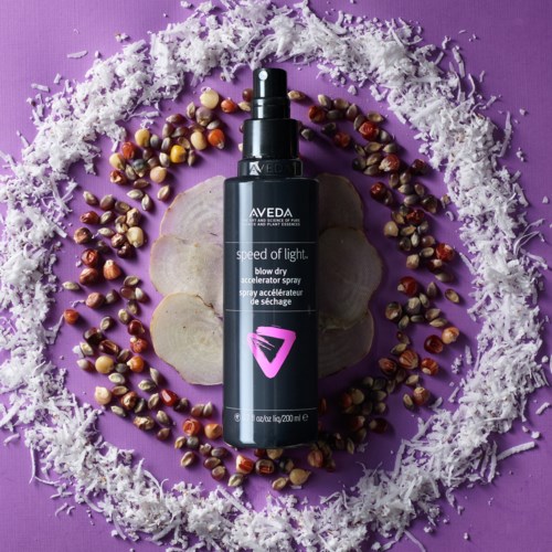 Musta suihkepullo, jonka etiketissä lukee 'Aveda', ympärillä siemeniä, kuoria ja valkoista raapaleita violetilla taustalla.
