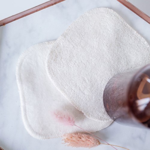 Katoenen pads en een bruine fles op een lichte achtergrond. Een katoenen pad ligt voor, met wat roze vezels in de buurt.