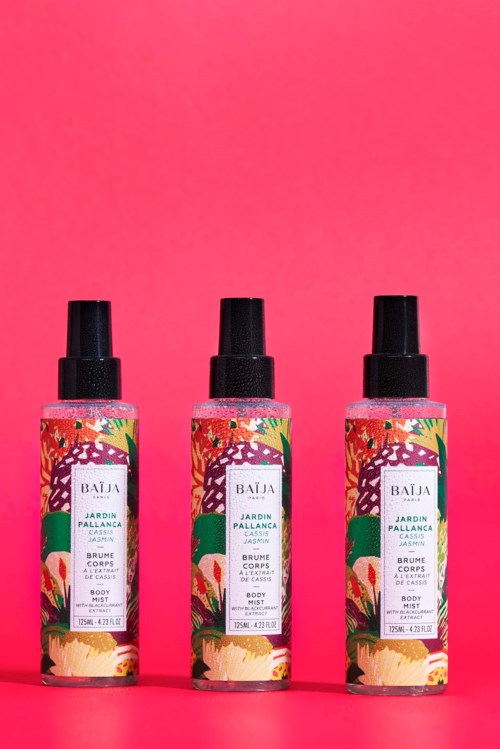 Trzy butelki spray do ciała z kolorowymi etykietami z kwiatami na różowym tle.