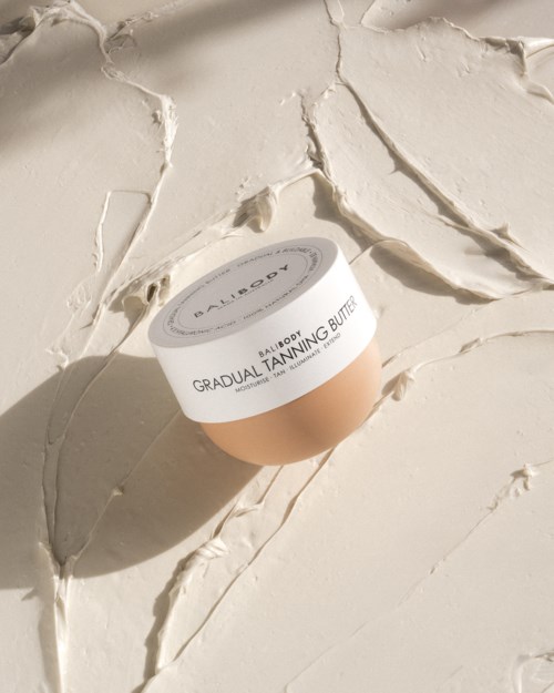 Ein Kosmetikbehälter mit 'Gradual Tanning Butter' liegt auf einer texturierten cremefarbenen Oberfläche.