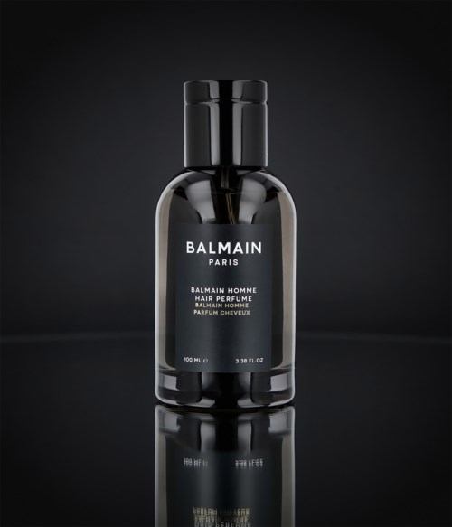 En svart flaske med en elegant etikett som sier 'Balmain Paris' og 'Hair Perfume', står mot en mørk bakgrunn.