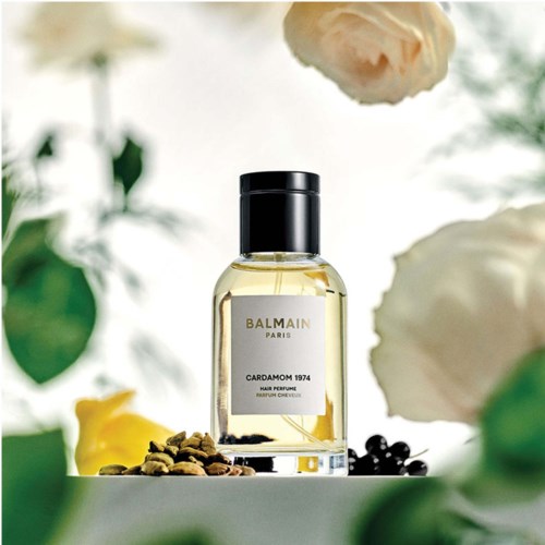 En parfyllflaska märkt 'Balmain Paris Cardamom 1974' omgiven av blommor och kryddor på en vit yta.